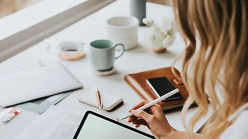 Mujer tomando notas en una tableta, con el móvil y una taza sobre el escritorio